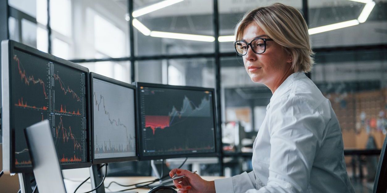 Female financial professional in an office looking at monitors displaying investment performance data.